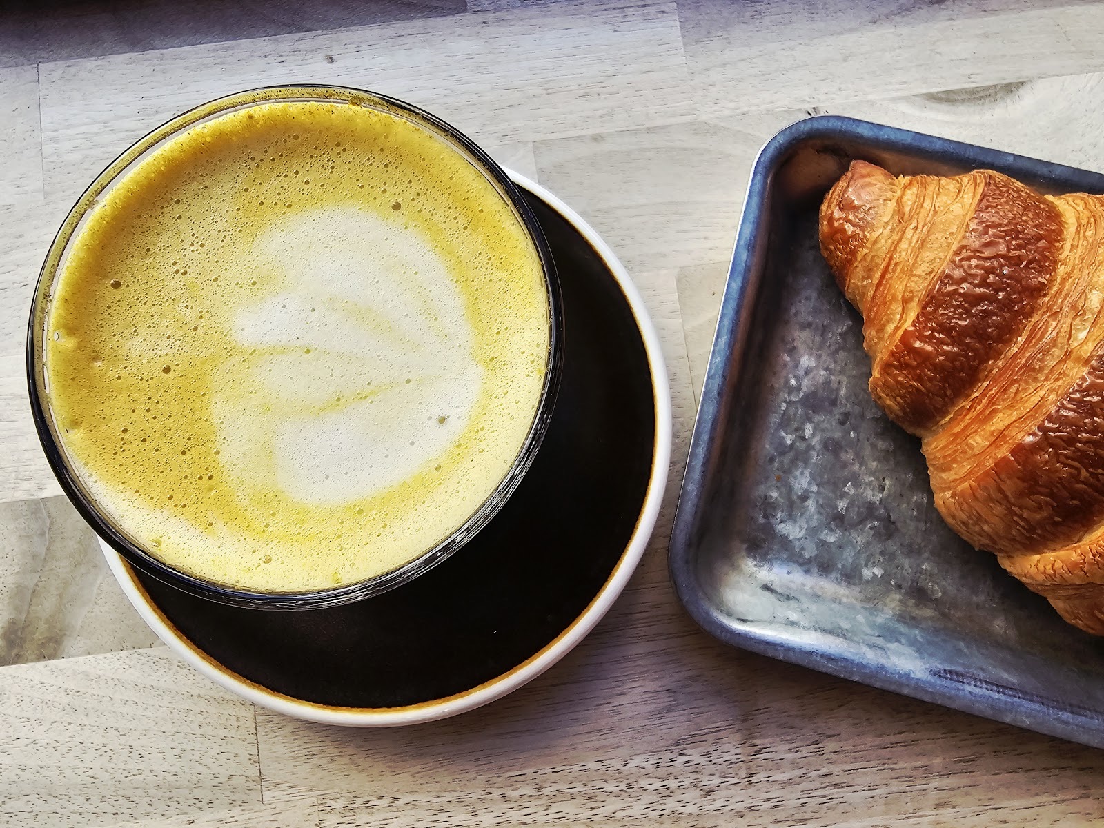 Flat white with leaf latte art beside a croissant on a metal tray